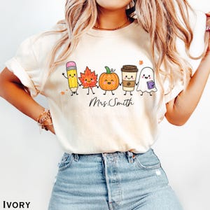 May include: An ivory t-shirt featuring a graphic of a pencil, a maple leaf, a pumpkin, a coffee cup, and a ghost holding hands. Each character has a smiling face. The text "Mrs. Smith" is printed in black cursive script below the characters.