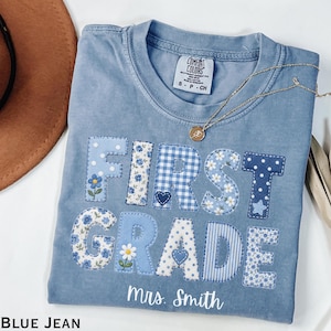 May include: A blue short-sleeve t-shirt with the text FIRST GRADE appliqued in various blue and white patterned fabrics including gingham, floral, and polka dots. The text Mrs. Smith is printed in white below the applique. A gold necklace is visible.