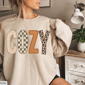 May include: A sand-colored crewneck sweatshirt featuring the word COZY printed across the chest. Each letter has a unique texture: the C is cream cable knit, the O is a green and white checkerboard, the Z is tan sherpa, and the Y is leopard print.