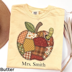 May include: A butter-colored t-shirt featuring a large apple graphic composed of various autumn-themed fabric patterns, including plaid, polka dots, and floral prints. A small green caterpillar with glasses sits on the apple. The text Mrs. Smith is printed below.