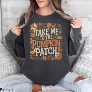 May include: Dark gray sweatshirt with the text "TAKE ME TO THE PUMPKIN PATCH" in orange and cream lettering. The design includes orange pumpkins and flowers in shades of orange, cream, and green. A fall-themed graphic.