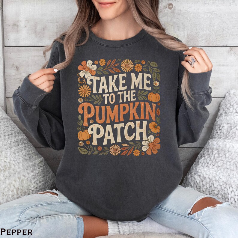 May include: Dark gray sweatshirt with the text "TAKE ME TO THE PUMPKIN PATCH" in orange and cream lettering. The design includes orange pumpkins and flowers in shades of orange, cream, and green. A fall-themed graphic.