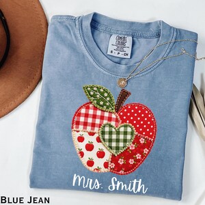 May include: A blue jean coloured t-shirt featuring a large apple-shaped applique made of various red, white, and green patterned fabric patches. The apple design includes a central green gingham heart and the white text Mrs. Smith printed below.