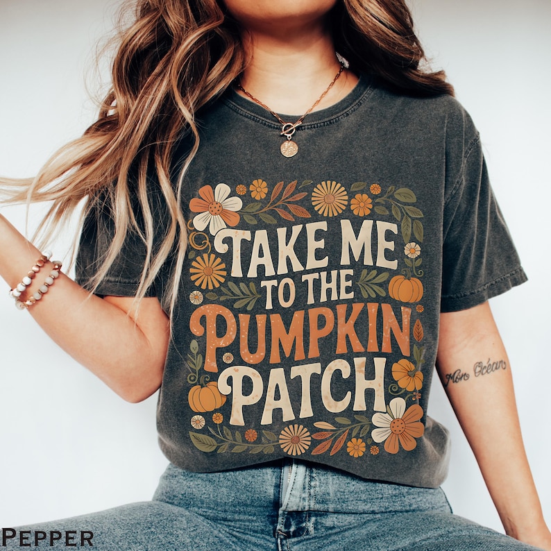 May include: Dark gray t-shirt with the text "TAKE ME TO THE PUMPKIN PATCH" in white and orange, surrounded by floral and pumpkin graphics. The shirt is a vintage style, perfect for fall.