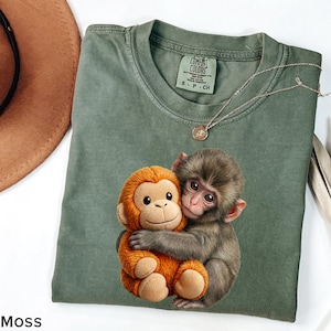 May include: Moss green Comfort Colors t-shirt featuring a graphic of two monkeys hugging. The shirt has a crew neck and short sleeves. A silver necklace with a small pendant is also visible. The shirt is labeled size S-P-CH.