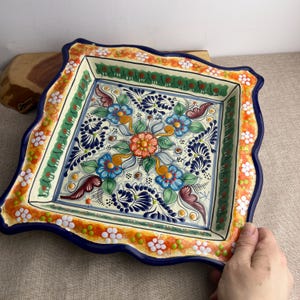 May include: Square ceramic serving dish with a scalloped edge. The dish features a vibrant floral design in blue, orange, green, and white. The rim is a deep blue, and the overall style is reminiscent of traditional hand-painted pottery.