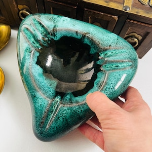 May include: A vintage ashtray with a distinctive shape and a glossy, teal glaze. The ashtray has a dark interior and a textured surface. It features grooves for cigarettes. A hand is holding the ashtray.