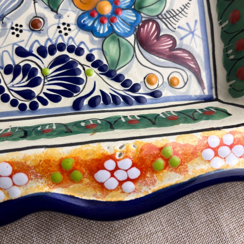 May include: A square, hand-painted ceramic serving dish with a vibrant floral design. The dish features a white base with blue, orange, green, and purple floral patterns. The rim is a deep blue with a scalloped edge.