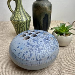 May include: A blue ceramic flower frog with speckled detailing and multiple holes for arranging stems. The frog is accompanied by a small potted succulent and two decorative vases in green and black.