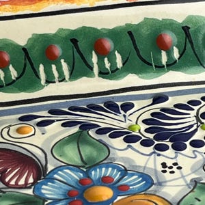 May include: Close-up of a decorative ceramic plate with a colorful floral design. The plate features a green border with red accents, blue and white floral patterns, and a vibrant color palette.