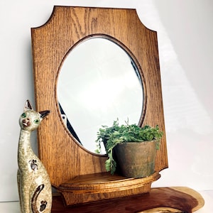 May include: A rectangular oak wood wall mirror with a central oval glass pane and a small integrated shelf at the base. A ceramic cat figurine with green eyes and brown paw print details stands to the left of the mirror, which holds a small plant.