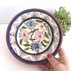May include: Hand-painted ceramic plate with a floral design. The plate has a blue rim and a decorative border with pink and blue accents. The centre of the plate showcases pink and blue flowers with green leaves.