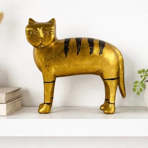 May include: A gold-coloured cat ornament with black stripes and details. The decorative cat statue is on a white shelf. The cat has a stylized design and is made of a metallic material.