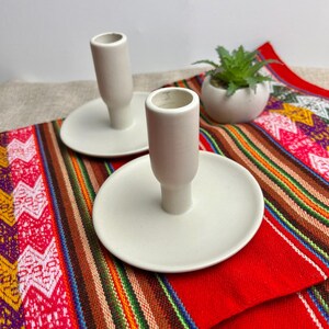 Peut inclure: Deux bougeoirs en céramique blanche au design simple. Chaque bougeoir a une base ronde et un corps cylindrique. Les bougeoirs sont posés sur des soucoupes blanches.