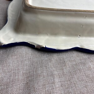 May include: A rectangular ceramic serving dish with a wavy edge. The dish has a white interior and a blue border. The edge of the dish has a wavy design. The dish is on a neutral-colored surface.
