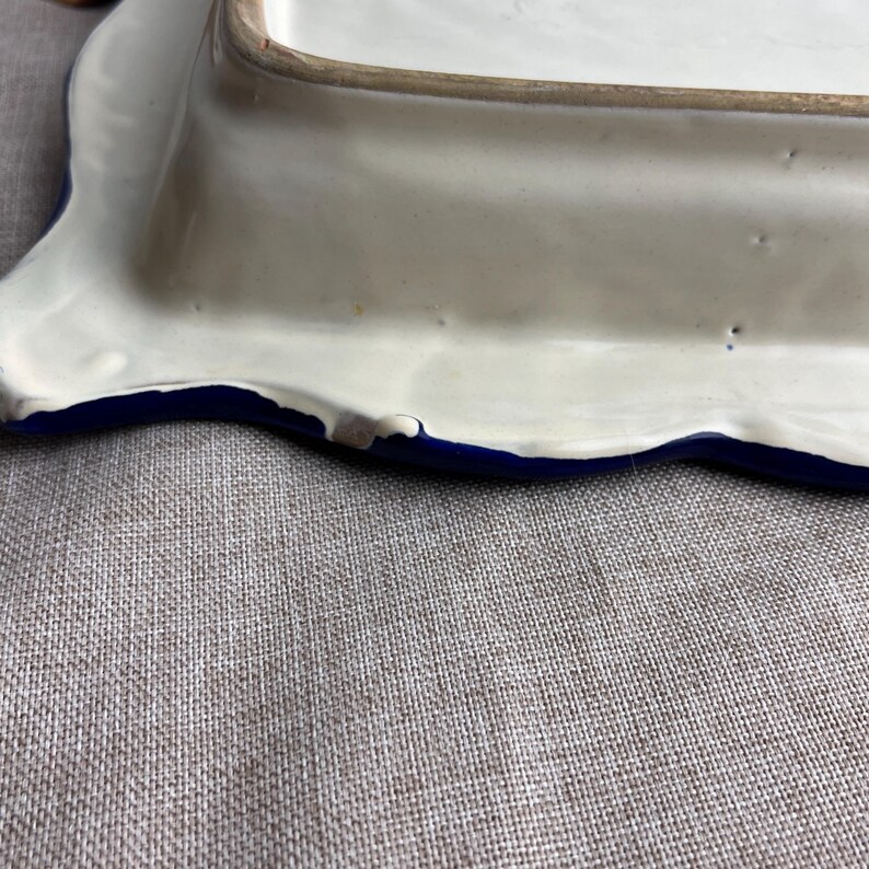 May include: A rectangular ceramic serving dish with a wavy edge. The dish has a white interior and a blue border. The edge of the dish has a wavy design. The dish is on a neutral-colored surface.