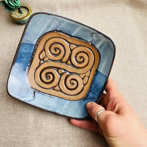 May include: Square ceramic plate with a blue and brown swirl design. The plate has a raised, textured pattern in the centre and a blue border. The plate is held by a hand.