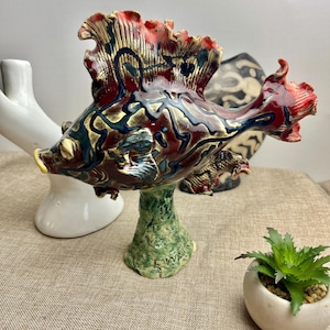 May include: A ceramic sculpture of a red, gold and blue fish with a green base. The fish has a large, flowing fin and a detailed, textured surface.