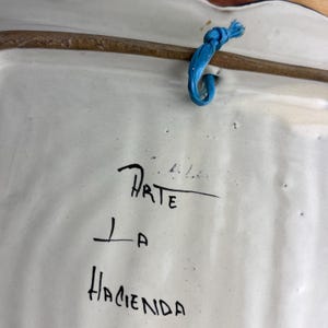 May include: Back of a white ceramic decorative plate with a brown rim and a blue string for hanging. The plate has the words "Arte La Hacienda" written in black ink.