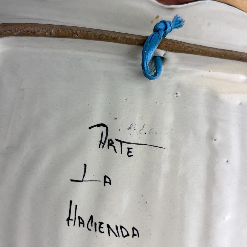 May include: Back of a white ceramic decorative plate with a brown rim and a blue string for hanging. The plate has the words "Arte La Hacienda" written in black ink.