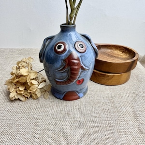 May include: Blue ceramic elephant-shaped vase with large eyes and a red heart on the snout. The vase is decorated with a brown elephant design and has a rustic, handmade look.