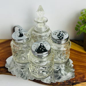 May include: A vintage glass condiment set on a silver-coloured metal tray. The set includes a cruet with a stopper, two shakers, and a lidded jar. The glass pieces have a diamond pattern.