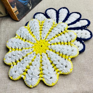 May include: Two daisy-shaped crochet coasters. The coasters are white with yellow centres and edges, and a dark blue border. They are made from yarn and are designed to protect surfaces from hot or cold drinks. Each coaster is approximately 15 cm in diameter.
