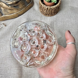 May include: A clear glass flower frog with multiple circular openings. The frog is round and held in a hand, with a silver ring on a finger. The background includes a woven basket with a small plant and a gold-coloured decorative object.