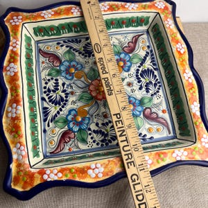 May include: Square ceramic serving dish with a hand-painted floral design. The dish features a blue, orange, green, and white color palette. The rim has a scalloped edge. The dish measures approximately 12 inches across.