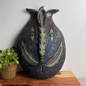 Peut inclure: Un masque mural en céramique bleu foncé texturé avec un visage stylisé. Comprend des yeux verts, des détails en forme de plumes et un petit accent orange. Le masque a un design artistique unique.