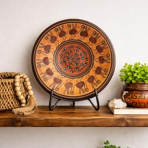 May include: A decorative ceramic plate with a brown rim and a tan centre, featuring a repeating pattern of stylized figures in brown and orange. The plate is displayed on a black metal stand on a wooden shelf.