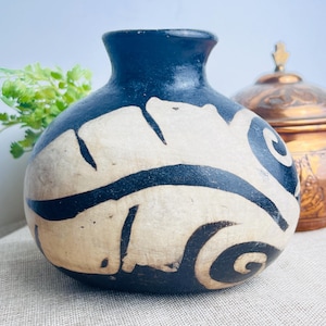 Peut inclure: Petit vase bleu foncé fait à la main, avec un corps arrondi et un col étroit. Le vase présente un motif abstrait crème avec des accents noirs. Un récipient décoratif en cuivre est visible en arrière-plan.