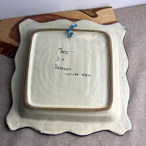 May include: Square ceramic plate with a scalloped edge, featuring a white base and a blue border. The back of the plate has handwritten text that reads "Arte La Hacienda Tlaxcala Mex." A blue string is attached for hanging.