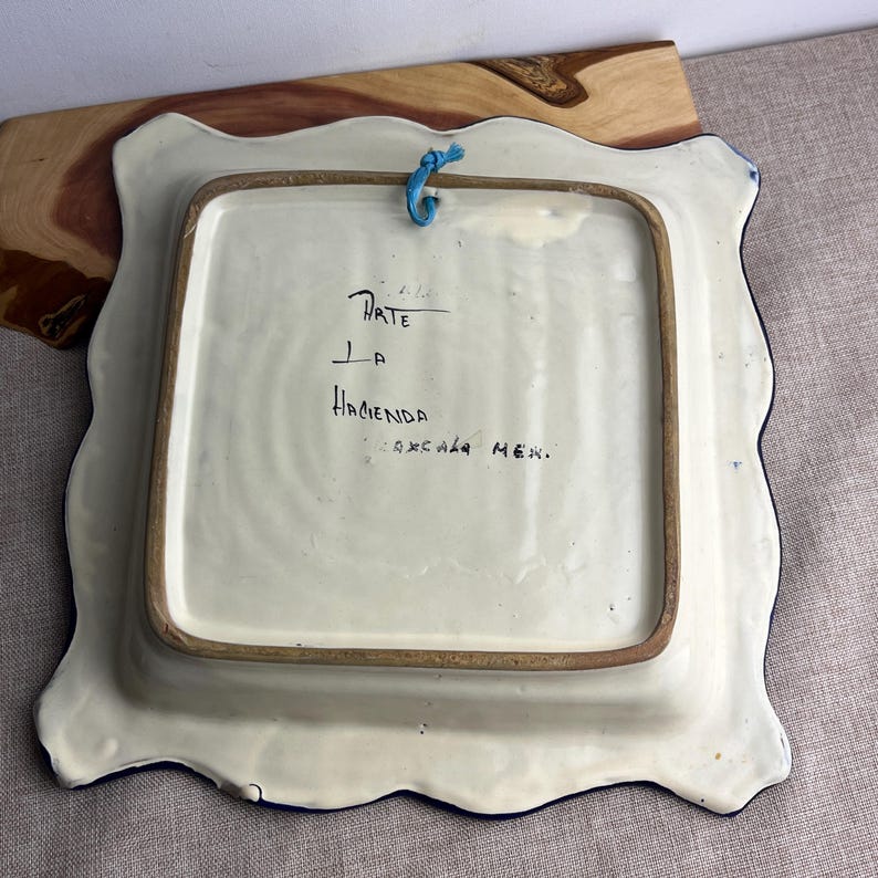 May include: Square ceramic plate with a scalloped edge, featuring a white base and a blue border. The back of the plate has handwritten text that reads "Arte La Hacienda Tlaxcala Mex." A blue string is attached for hanging.