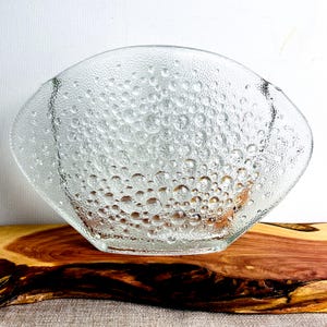 May include: Clear glass bowl with a textured, bubble-like pattern. The bowl has a wide, shallow shape and sits on a wooden surface. The glass bowl is a decorative piece.
