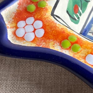 May include: Close-up of a ceramic dish with a vibrant design. The dish features a deep blue rim, an orange and yellow center, and white and green decorative elements. The design includes white flower-like shapes and green dots.