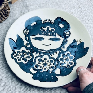 May include: A white ceramic plate featuring a blue and white illustration of a stylised woman with flowers. The plate is signed "LGT" in gold.