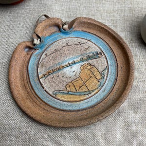 May include: A handmade ceramic wall hanging with a coastal scene. The plate has a brown rim, a blue inner ring, and a central design with a bridge, buildings, and a rock formation. It includes a loop for hanging.