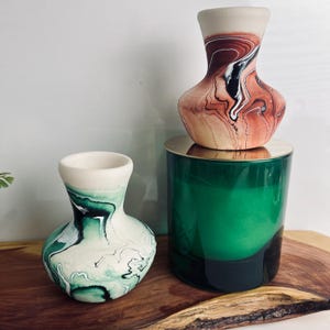 May include: Two small ceramic vases with marbled patterns and a large green glass candle. One vase features swirls of terracotta, black, and white, while the other displays shades of green, black, and white. The green candle has a gold lid.