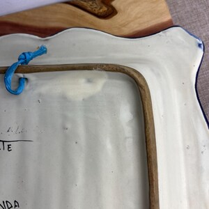 May include: A white ceramic serving platter with a scalloped edge and a blue border. The back of the platter has a brown frame and a blue string for hanging. The platter has some writing on it.