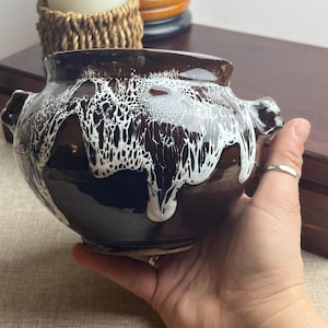 May include: A brown ceramic pot with white dripped glaze. The pot has two small handles on either side. The glaze creates a unique pattern across the surface. The pot is likely used for decorative or functional purposes.