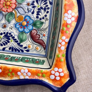 May include: A square ceramic serving dish with a vibrant floral design. The dish features a white base with blue, orange, red, and green floral patterns. The rim is decorated with white flower and green dot accents, framed by a blue border.