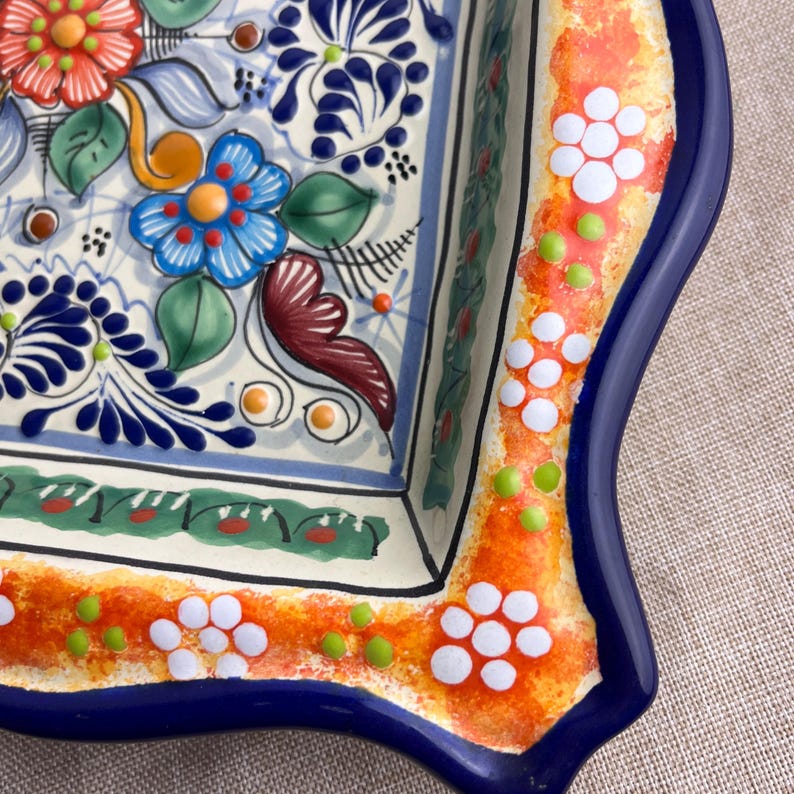 May include: A square ceramic serving dish with a vibrant floral design. The dish features a white base with blue, orange, red, and green floral patterns. The rim is decorated with white flower and green dot accents, framed by a blue border.