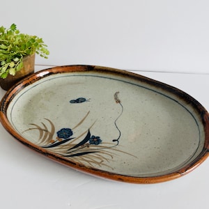 May include: Oval ceramic serving platter with a speckled, light grey surface and a brown rim. The platter is decorated with blue flowers, brown grass-like designs, and a small butterfly. A decorative piece for serving food.