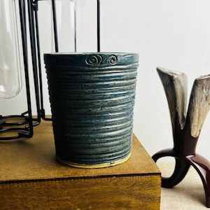 May include: A handmade ceramic tumbler with a textured, ribbed design in shades of blue and grey. The top rim features decorative swirl details. The base has a yellow-beige colour. The tumbler is displayed on a wooden surface.