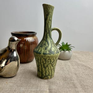 May include: A green ceramic vase with a textured surface and a handle. The vase has a narrow neck and a wide base.