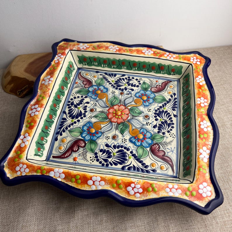 May include: Square ceramic serving dish with a scalloped edge. The dish features a floral design in blue, orange, green, and white. The rim is orange with white flower accents, and the edge is a deep blue.