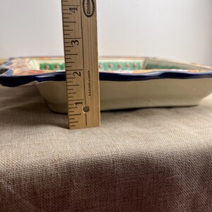 May include: A rectangular ceramic serving dish with a cream-colored base and a blue rim. The dish features a colorful floral design with orange, green, and white accents. The dish is approximately 2 inches (5 cm) deep.