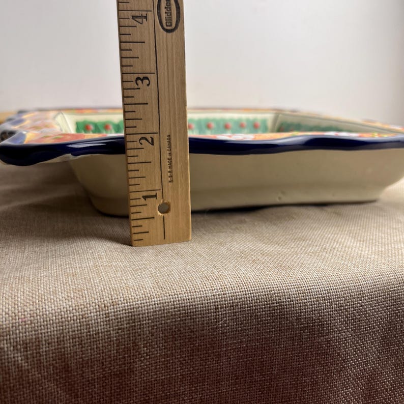 May include: A rectangular ceramic serving dish with a cream-colored base and a blue rim. The dish features a colorful floral design with orange, green, and white accents. The dish is approximately 2 inches (5 cm) deep.