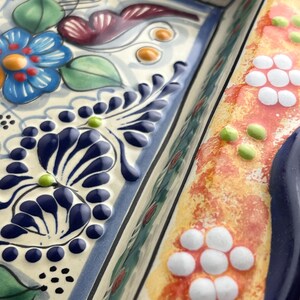 May include: Close-up of a decorative ceramic serving dish with a raised floral design. The dish features a blue flower, dark blue leaves, and white and orange accents. The edges are outlined in blue.